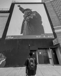 A young man stands with his back to the camera looking up at a billboard photograph of a Black journalist with one fist raised